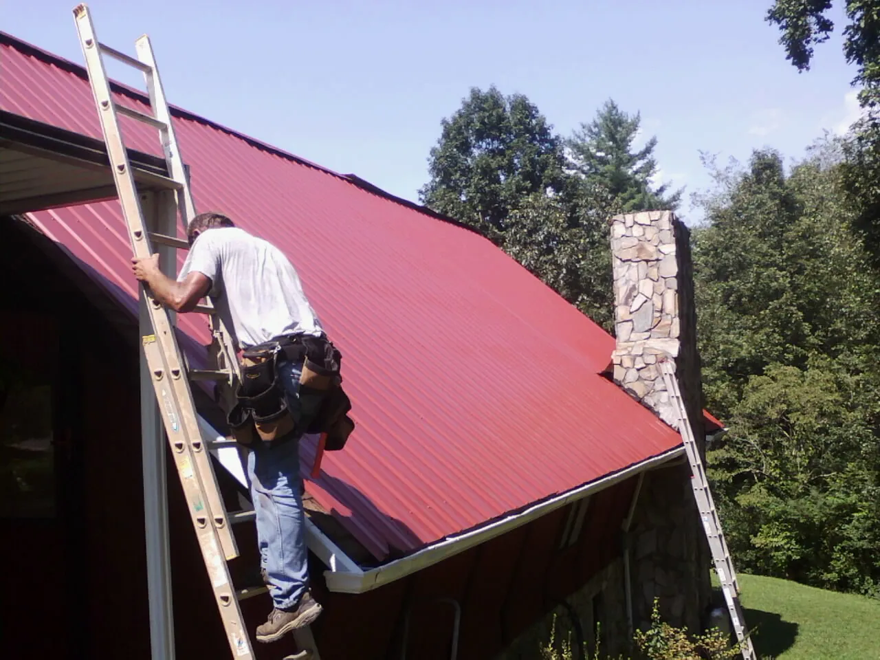 Roofer on a ladder beside a red metal roof and stone chimney — Soffit & Fascia Repair by Roofing Brothers in San Jacinto